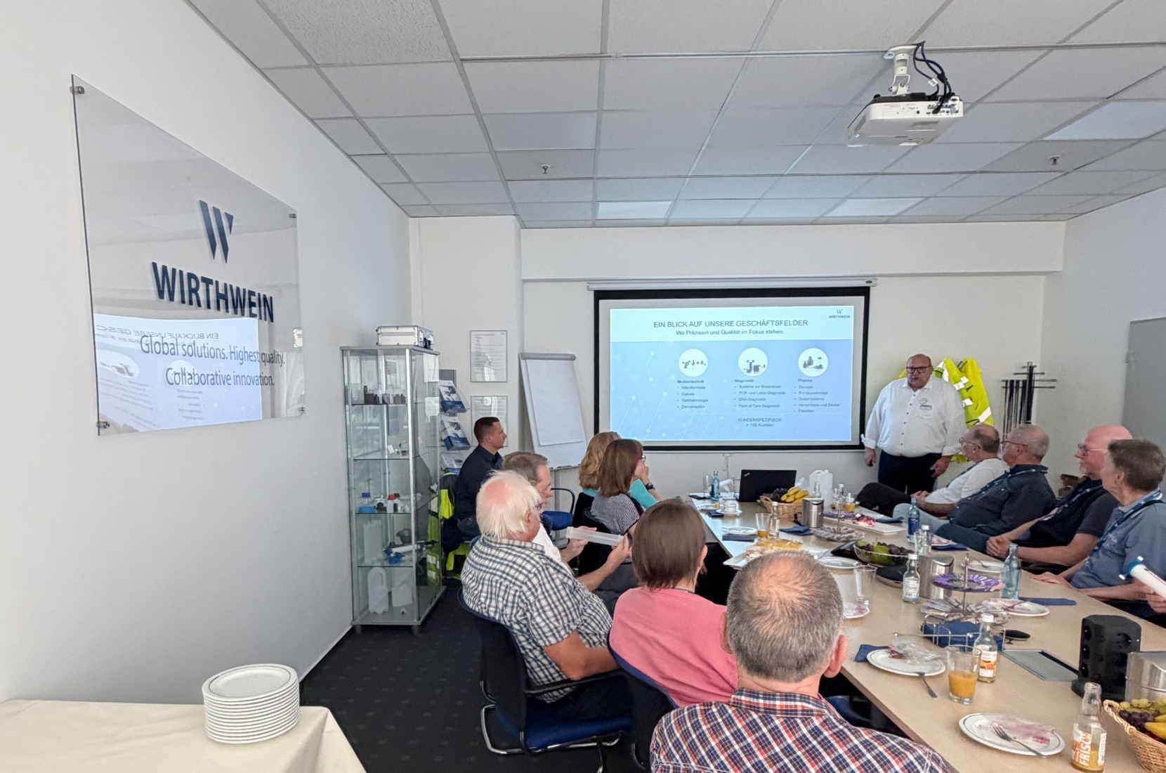 Mehrere Besucherinnen und Besucher sitzen an einem Besprechungstisch während einer Präsentation bei Wirthwein Medical. Eine Person steht vor einer Leinwand und erläutert Inhalte zu den Geschäftsfeldern des Unternehmens. Auf dem Tisch stehen Getränke, Obst und Informationsmaterialien. Im Hintergrund sind das Wirthwein-Logo, eine Vitrine sowie die Präsentationsfläche zu sehen. Text im Bild: „Tobias Müller, Leiter Administration und Logistik, gibt den Besucherinnen und Besuchern einen Einblick in Wirthwein Medical und die vielfältigen Einsatzmöglichkeiten von Kunststoff."