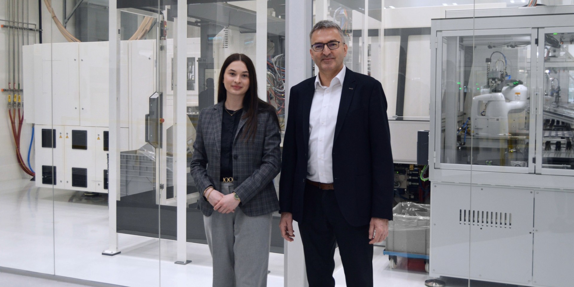 Werkstudentin Rosalina Bütün und Geschäftsführer Dr. Thomas Jakob stehen vor einer verglasten Reinraumanlage der Klasse 7 bei Wirthwein Medical. Im Hintergrund sind automatisierte Produktionsanlagen und technische Systeme des Reinraumbereichs zu sehen. Text im Bild: „Werkstudentin Rosalina Bütün gemeinsam mit Geschäftsführer Dr. Thomas Jakob vor dem Reinraum der Klasse 7."